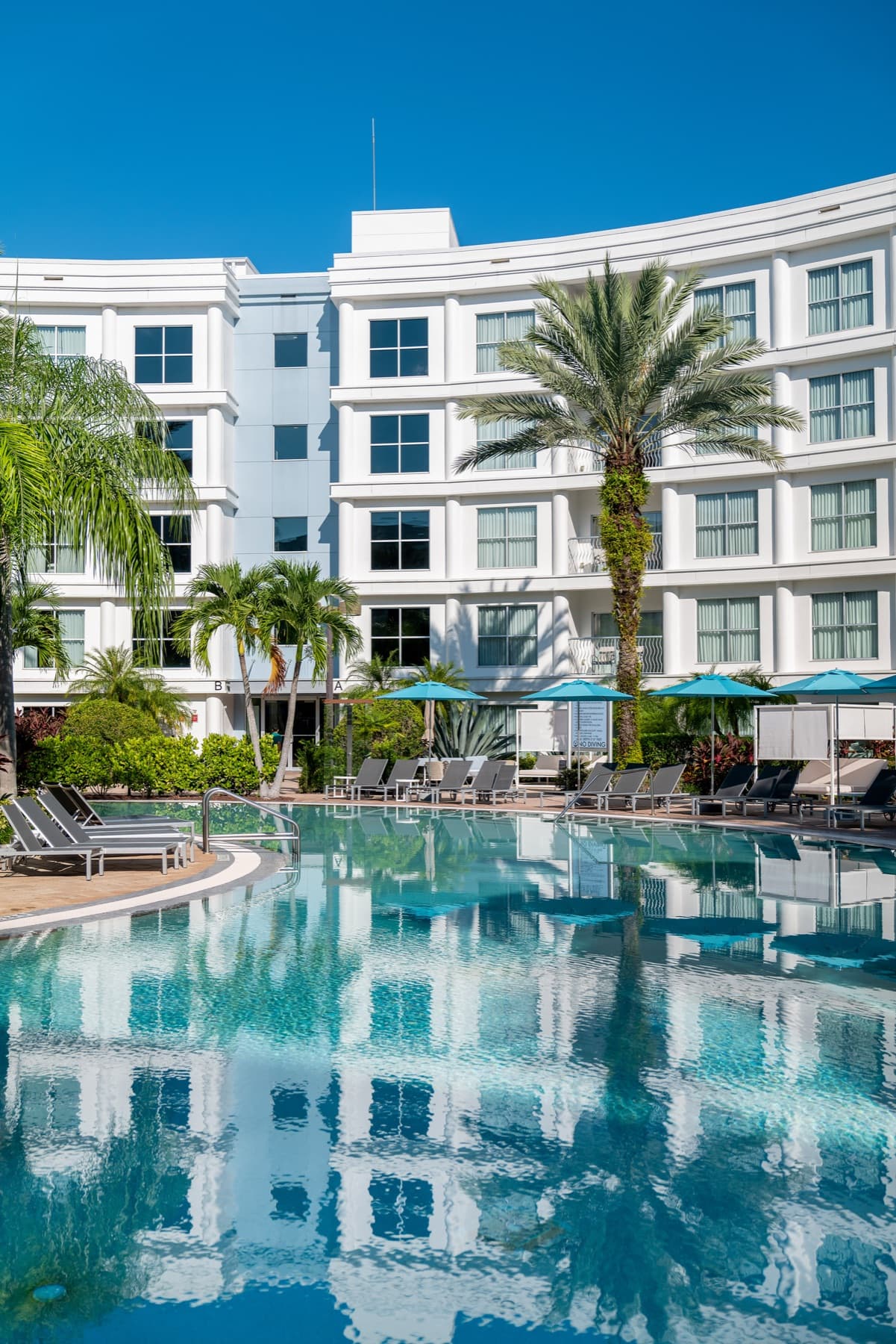 The curved white facade of Meliá Orlando reflected in its courtyard pool, beneath palms and teal parasols.