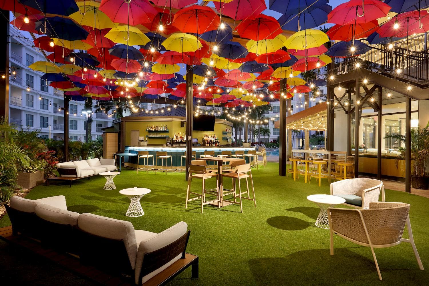 An outdoor hotel bar terrace at dusk beneath a canopy of coloured umbrellas and festoon lights.