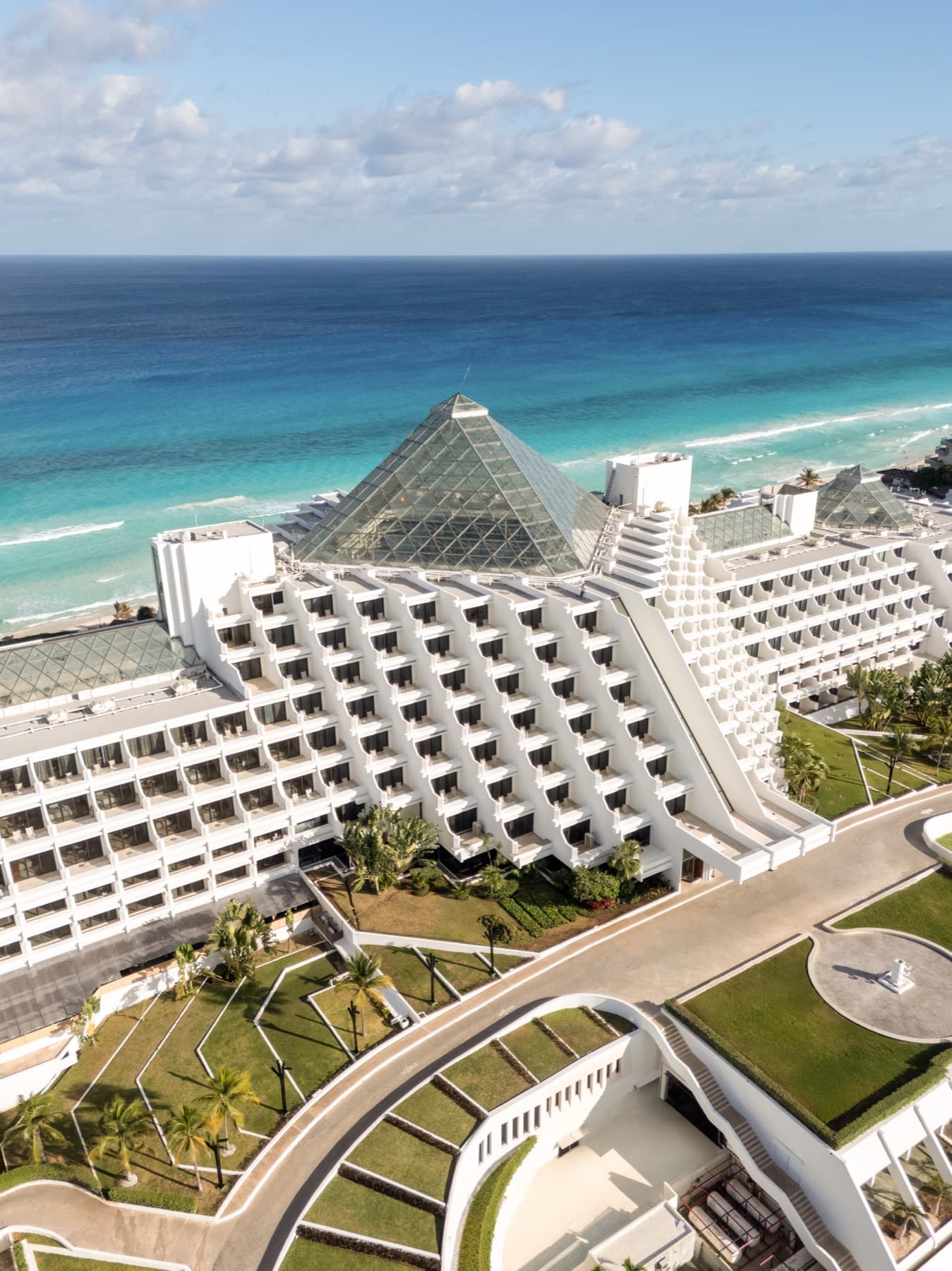Aerial view of Paradisus Cancún, its terraced white wings and glass pyramid atrium set above a turquoise sea.